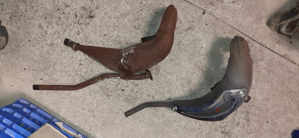 Dos bufandas de escape para ciclomotor sobre suelo de taller; una con óxido superficial y marcaje manual para Rieju RR 50 y otra con protector térmico cromado para Aprilia SR 50.