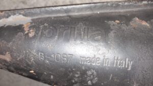 Primer plano del cuerpo metálico de un escape para ciclomotor Aprilia SR 50, donde se aprecia el grabado original en relieve con la inscripción "aprilia" y el código técnico "B-1087 made in italy".