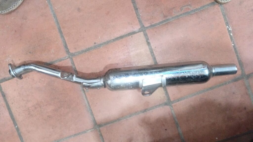 Silenciador de escape tipo custom cromado de diseño universal, visto desde el lateral, con soporte de anclaje y colector integrado sobre suelo de baldosa.