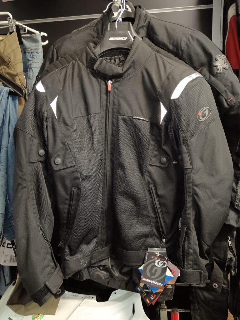 Chaqueta de moto de malla Garibaldi negra colgada en una tienda. Muestra paneles de ventilación, detalles reflectantes y etiquetas de marca.