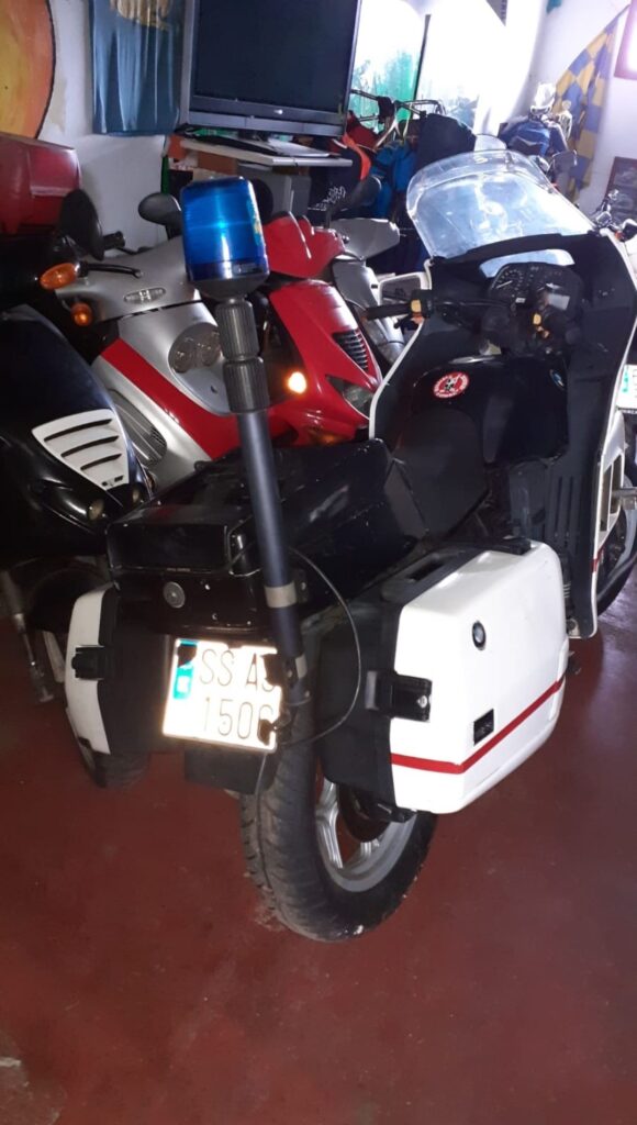 Vista trasera de moto BMW K75 de 1992 blanca estilo patrulla con maletas rígidas, mástil de sirena azul y matrícula.