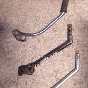 Tres pedales de arranque variados para motos clásicas sobre suelo de hormigón, con cajas azules de plástico al fondo.