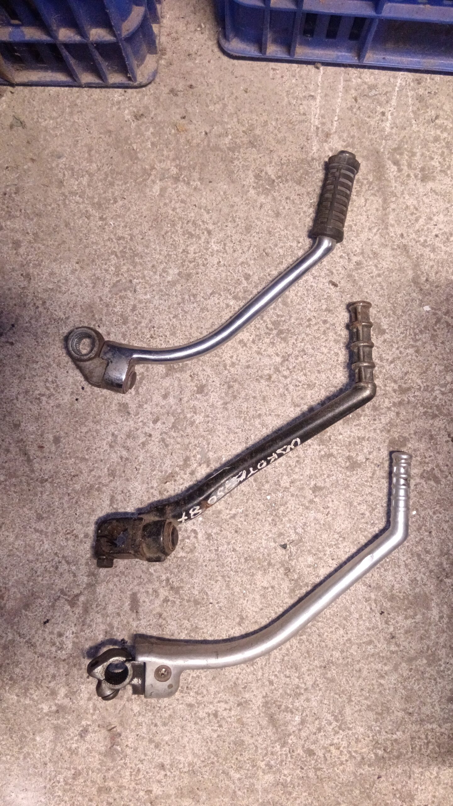 Tres pedales de arranque variados para motos clásicas sobre suelo de hormigón, con cajas azules de plástico al fondo.