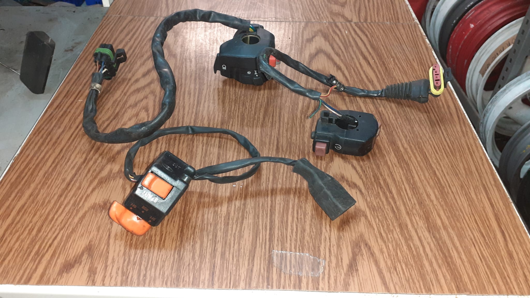 Mandos de manillar y conmutadores de luces para moto usados. Tres unidades con cableado y conectores visibles sobre madera.
