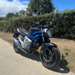 Vista frontal tres cuartos de moto naked Suzuki Gladius 650 azul y negra de 2010 aparcada en un camino asfaltado con sol.