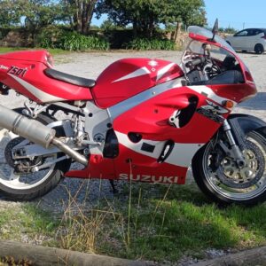 Perfil lateral derecho de una Suzuki GSX-R 750 SRAD de 1999 en color rojo y blanco. La moto luce su carenado original con inyección electrónica (750i), chasis de doble viga de aluminio y un escape deportivo de alto rendimiento, estacionada en un entorno exterior.