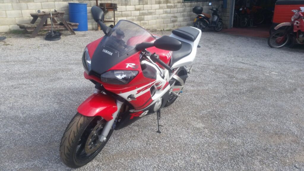 Moto deportiva Yamaha R6 de 1999 en color rojo y blanco, vista frontal de tres cuartos, aparcada en un recinto exterior.