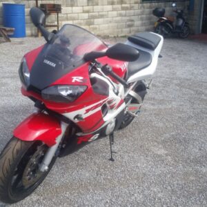 Moto deportiva Yamaha R6 de 1999 en color rojo y blanco, vista frontal de tres cuartos, aparcada en un recinto exterior.