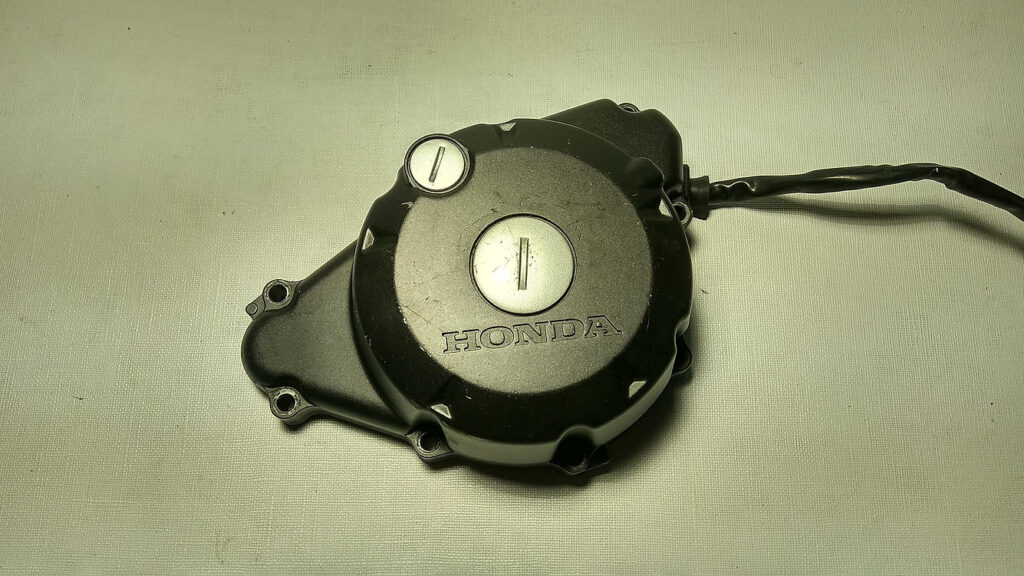Vista exterior de la tapa de alternador y cárter de encendido original negra con logotipo grabado para motocicleta Honda CBR 125 R años 2004 a 2006.