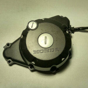 Vista exterior de la tapa de alternador y cárter de encendido original negra con logotipo grabado para motocicleta Honda CBR 125 R años 2004 a 2006.