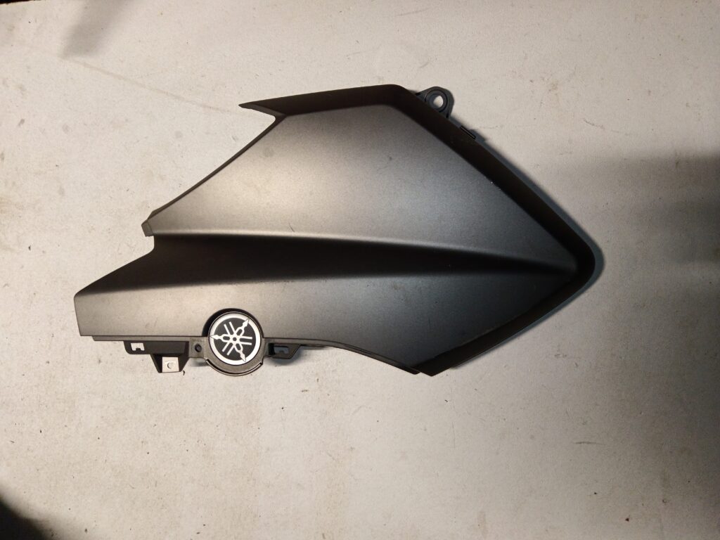 Tapa lateral izquierda negra original para Yamaha XMAX 125, vista exterior con emblema de la marca.